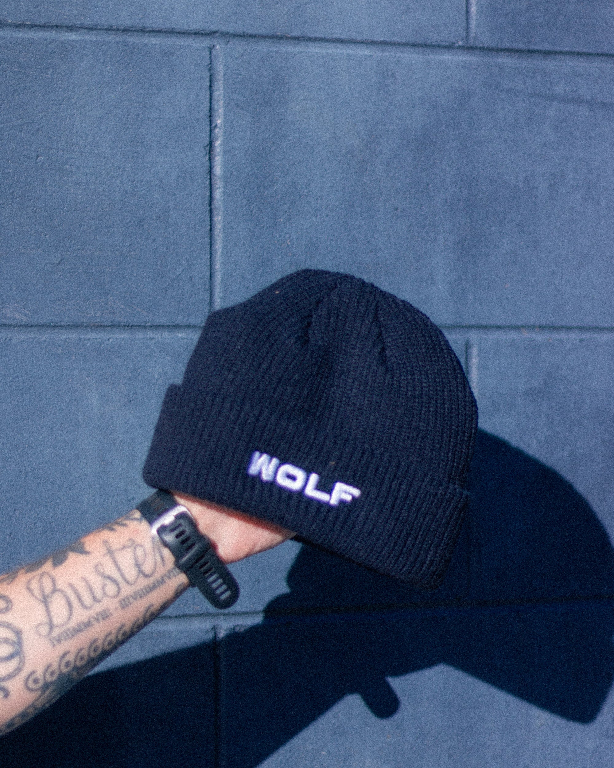 Navy beanie with 'WOLF' logo held against a blue tiled wall